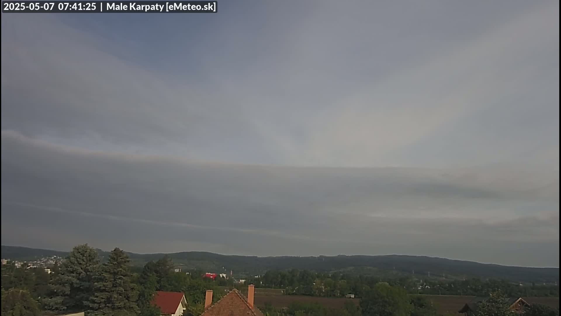 A mostly cloudy sky hangs over a hilly landscape dotted with trees and houses.