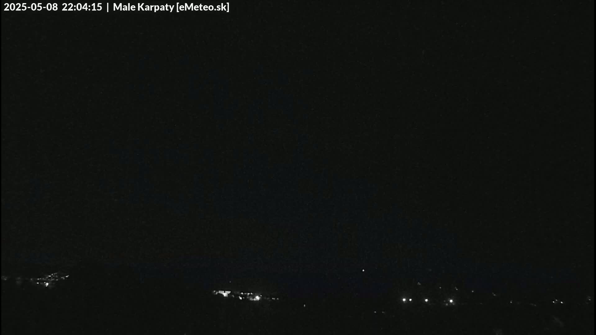 A nighttime, dark image shows a dimly lit landscape with a few small lights visible in the distance under a clear, dark sky.