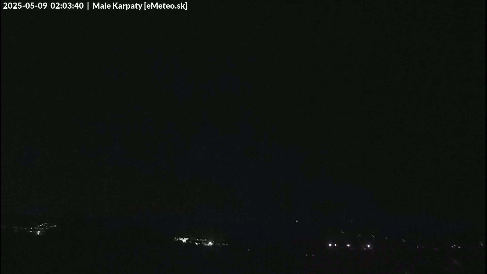 A dark nighttime landscape shows a few faint lights in the distance under a clear, black sky.