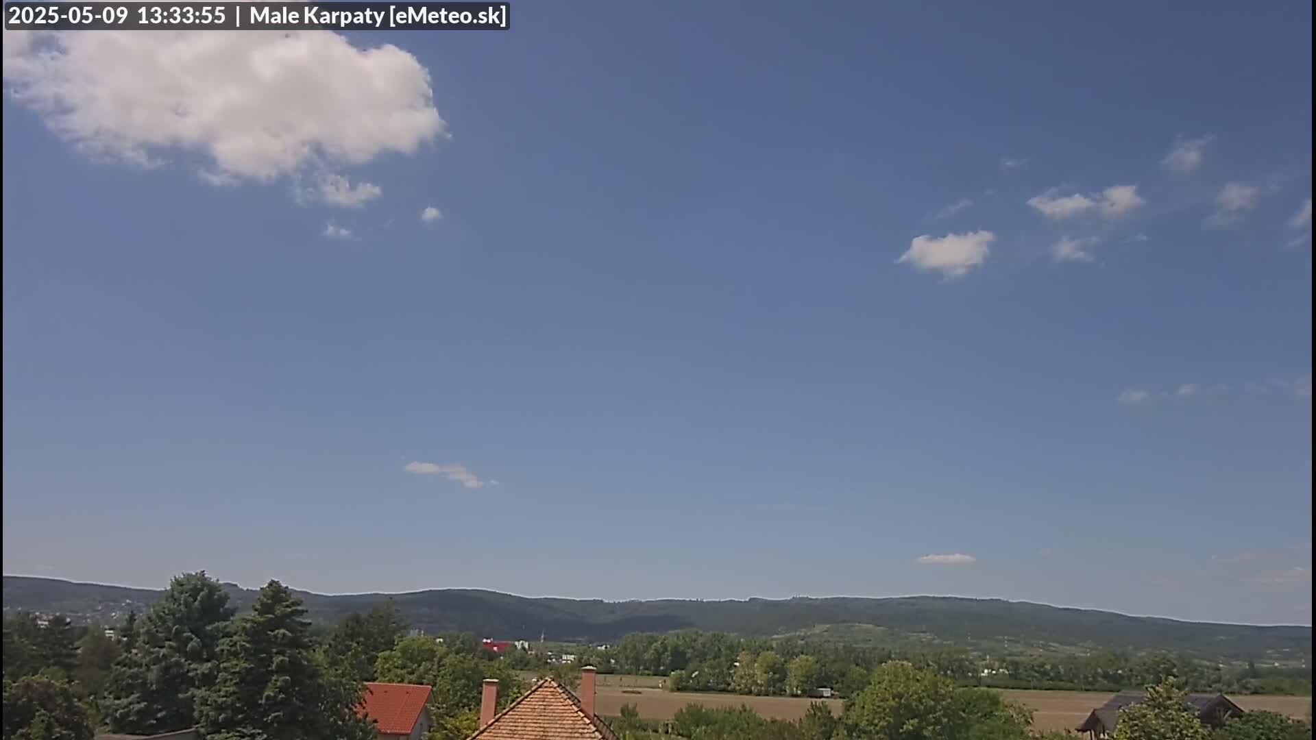 A mostly clear blue sky with a few scattered clouds overlooks a landscape of green trees and rolling hills.
