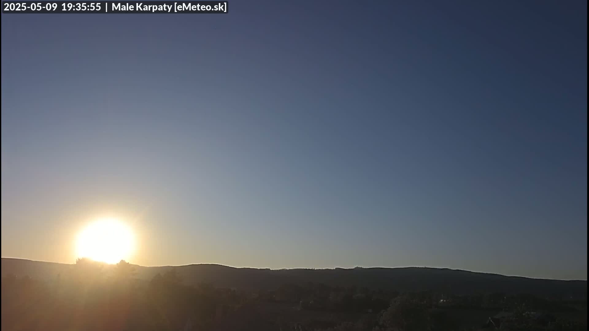 A bright sun sets behind a silhouetted line of hills under a clear, twilight sky.
