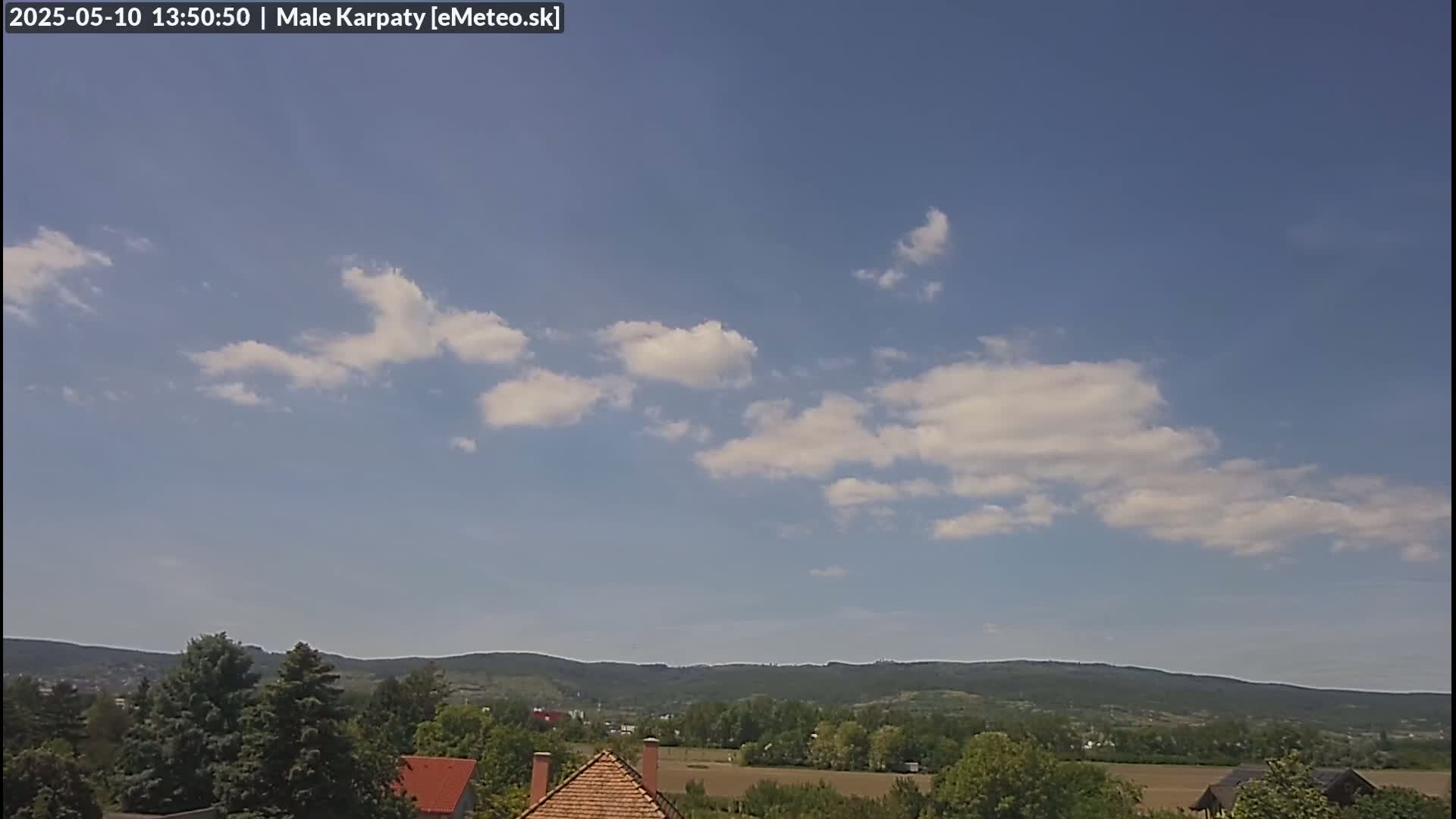 A mostly clear blue sky with scattered cumulus clouds overlooks a landscape of green hills and trees, with a few buildings visible in the foreground.
