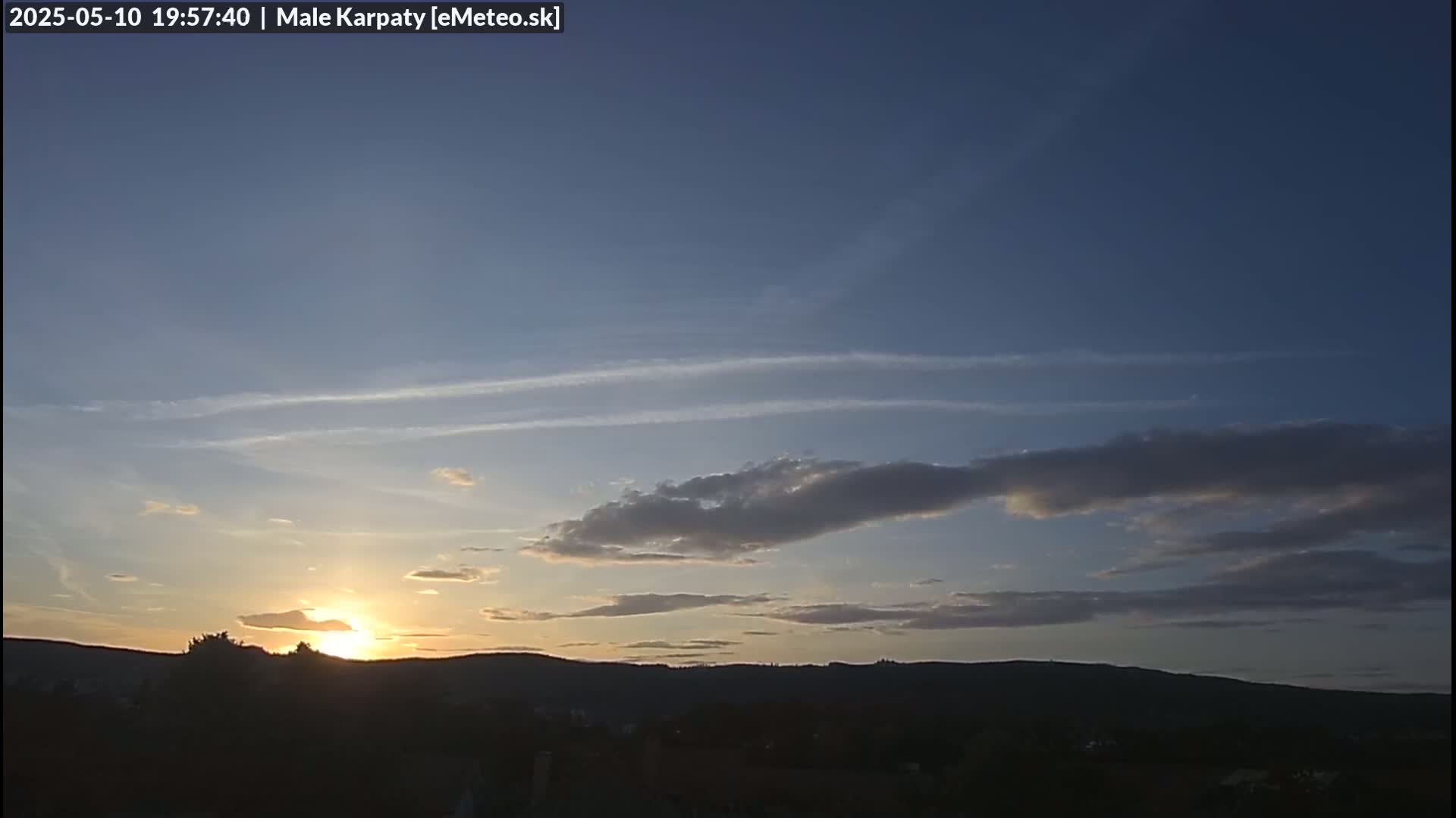 The setting sun casts a golden glow on a partly cloudy sky above a dark, silhouetted mountain range.