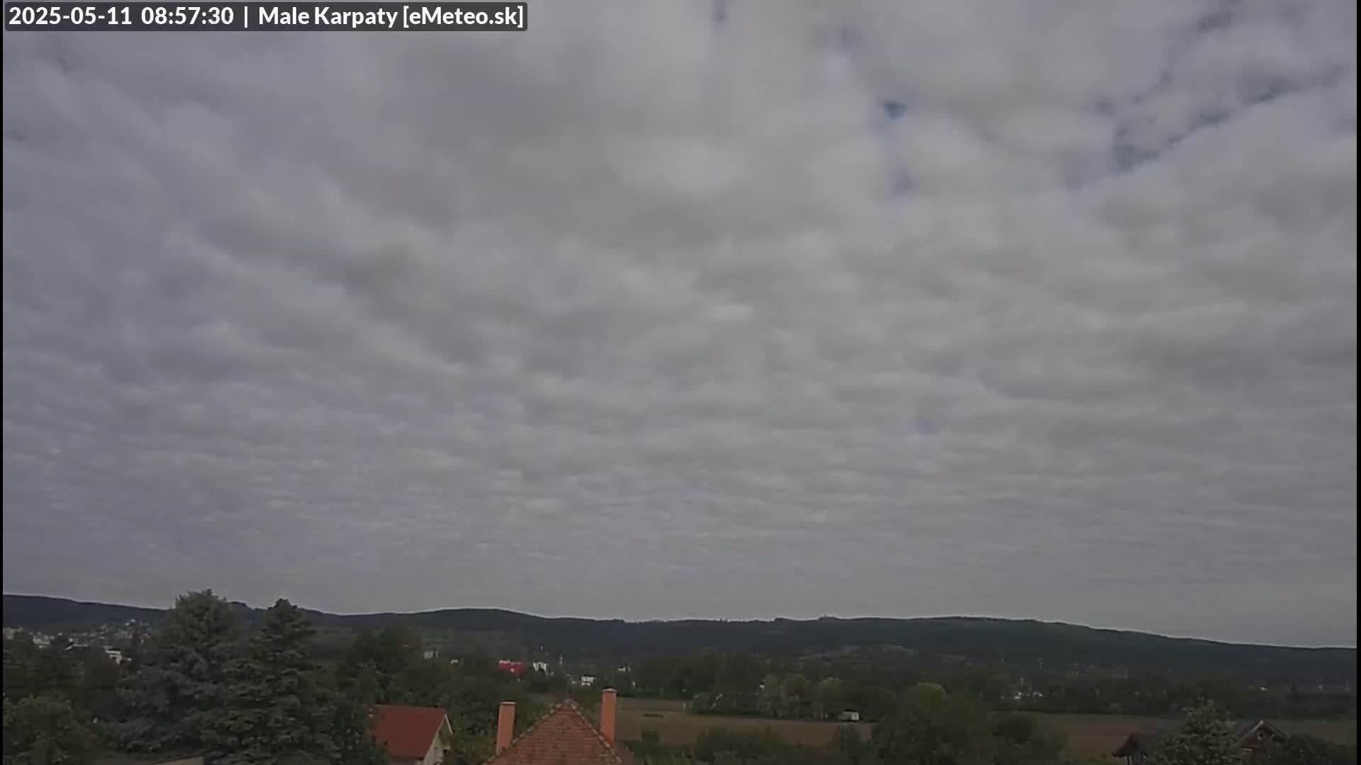 A mostly cloudy sky hangs over a landscape of green hills and trees, with a few houses visible in the foreground.