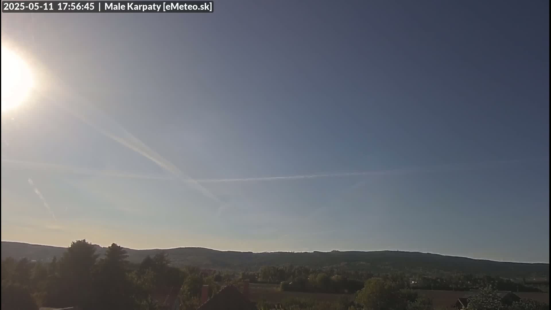 A bright sun shines in a mostly clear sky over a line of low hills covered in green vegetation.