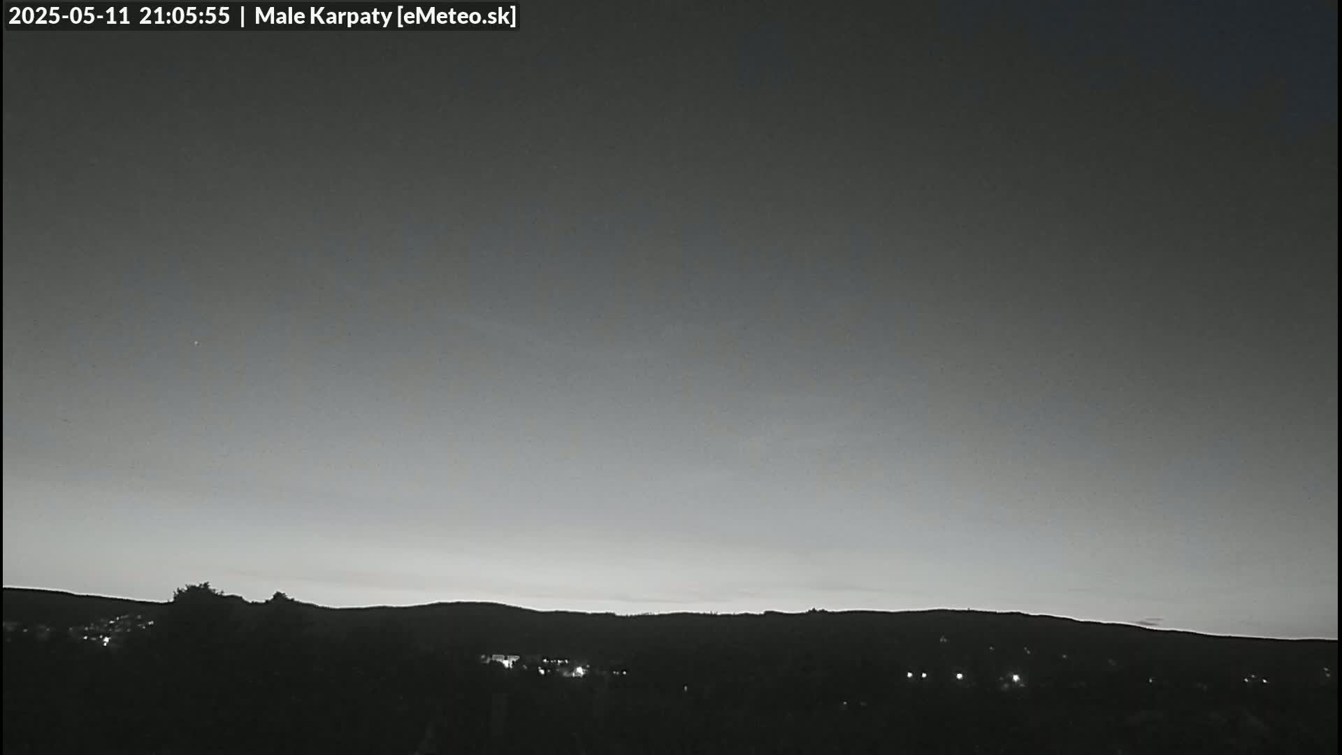 A grayscale image shows a dark, mostly clear night sky over a silhouetted landscape of hills and trees with a few faint lights visible in the distance.