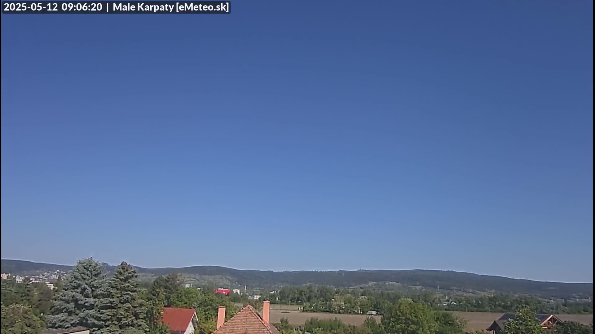 Under a clear blue sky, a view of houses and green trees stretches towards distant, low-lying mountains.