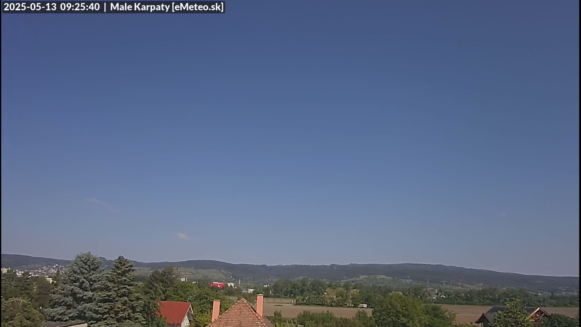 A clear blue sky overlooks a tree-lined landscape with distant mountains and a few houses with reddish-brown roofs.