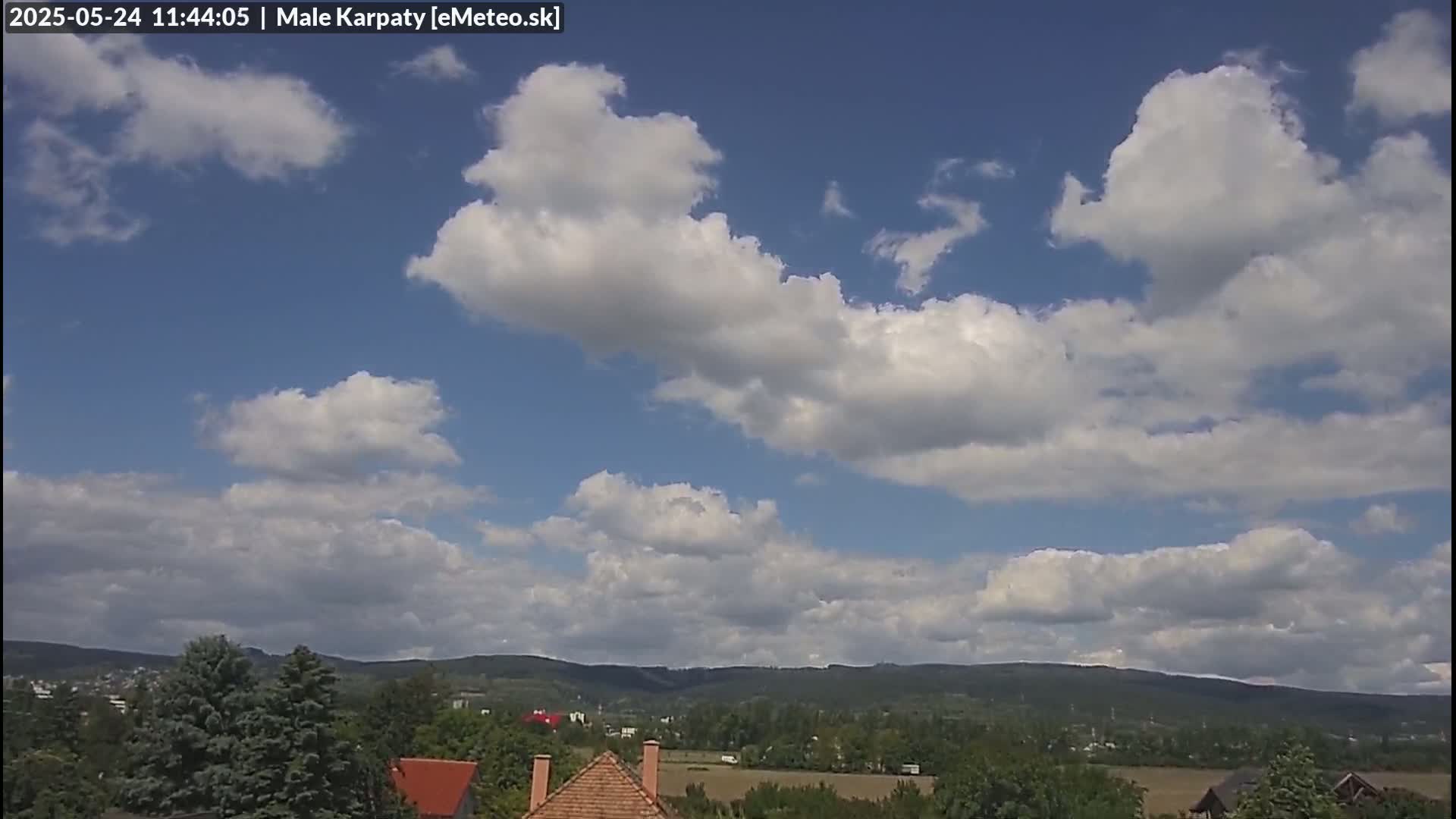 A partly cloudy sky with cumulus clouds over a landscape of rolling green hills and houses.