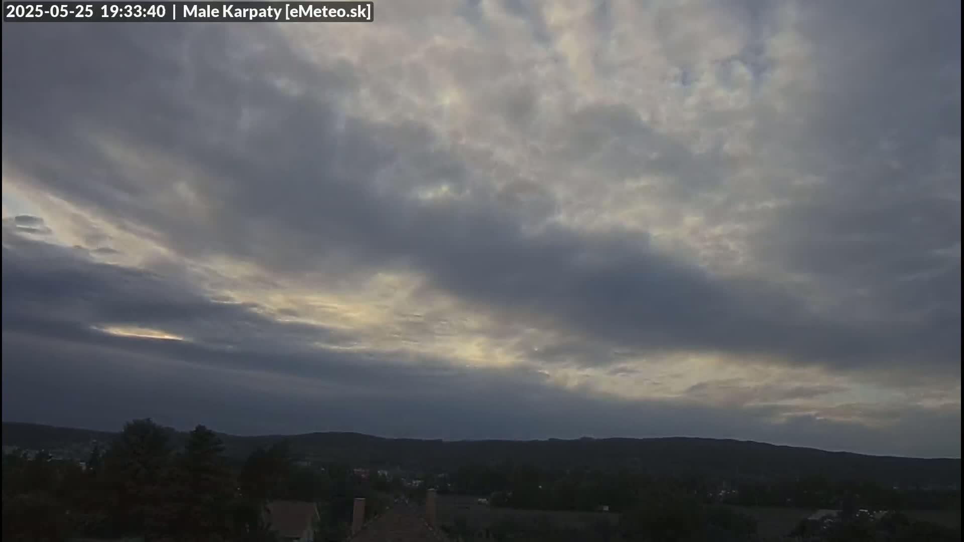 A mostly cloudy sky with some breaks of light showing through, above a dark, tree-lined hilly landscape.