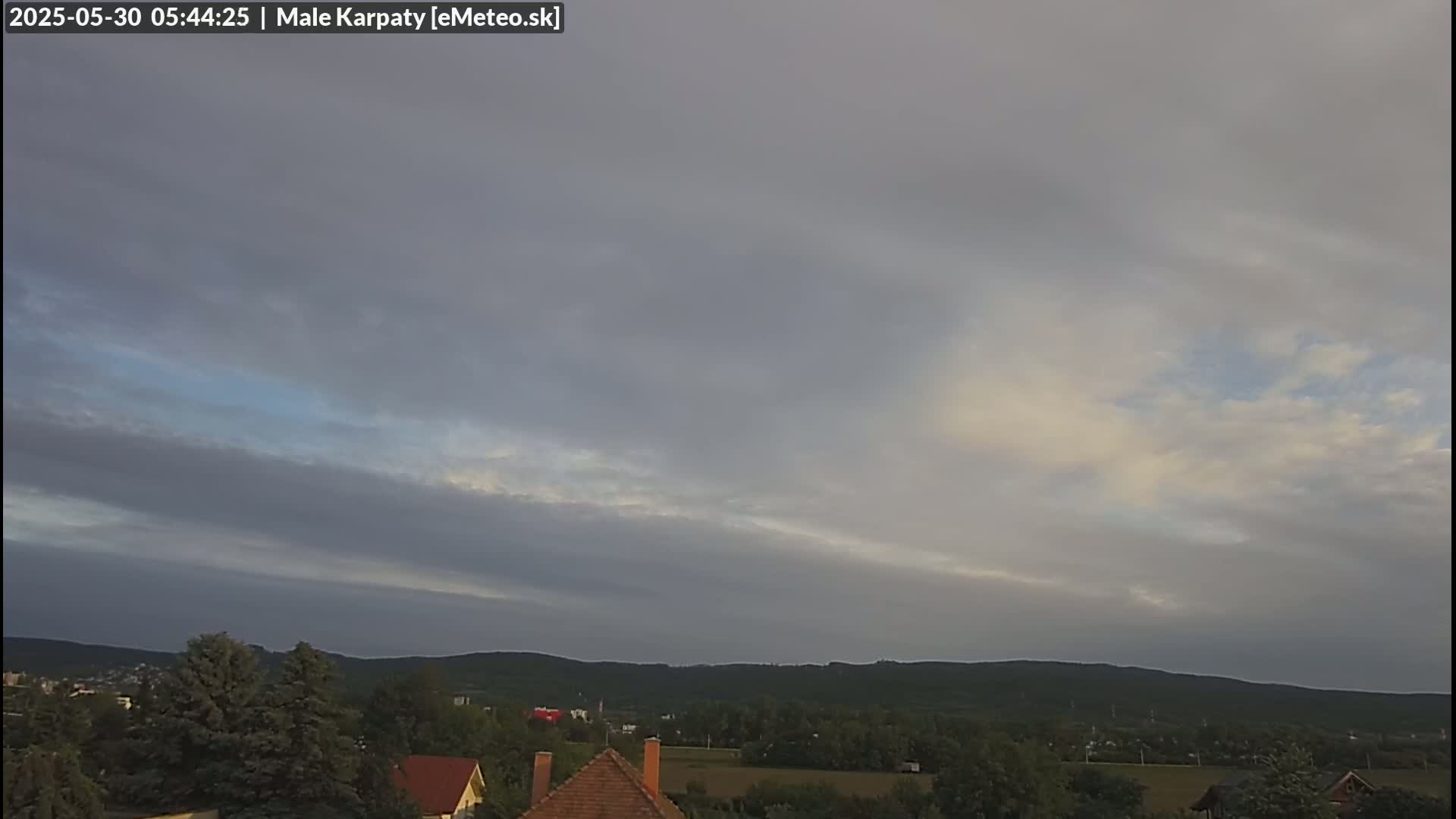 A mostly cloudy sky with hints of sunrise illuminates a distant, tree-covered mountain range overlooking a small town.