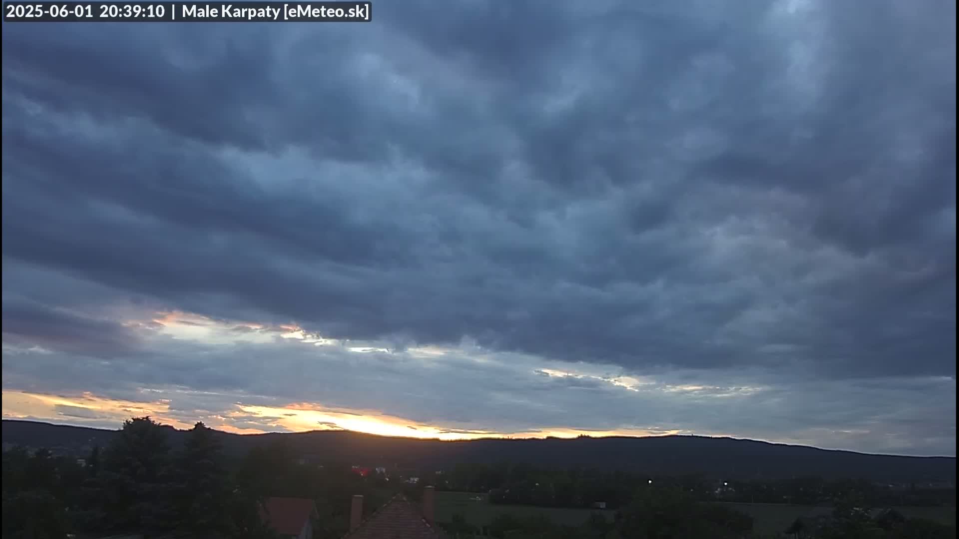 A mostly cloudy sunset over a distant mountain range with a few trees and buildings in the foreground.