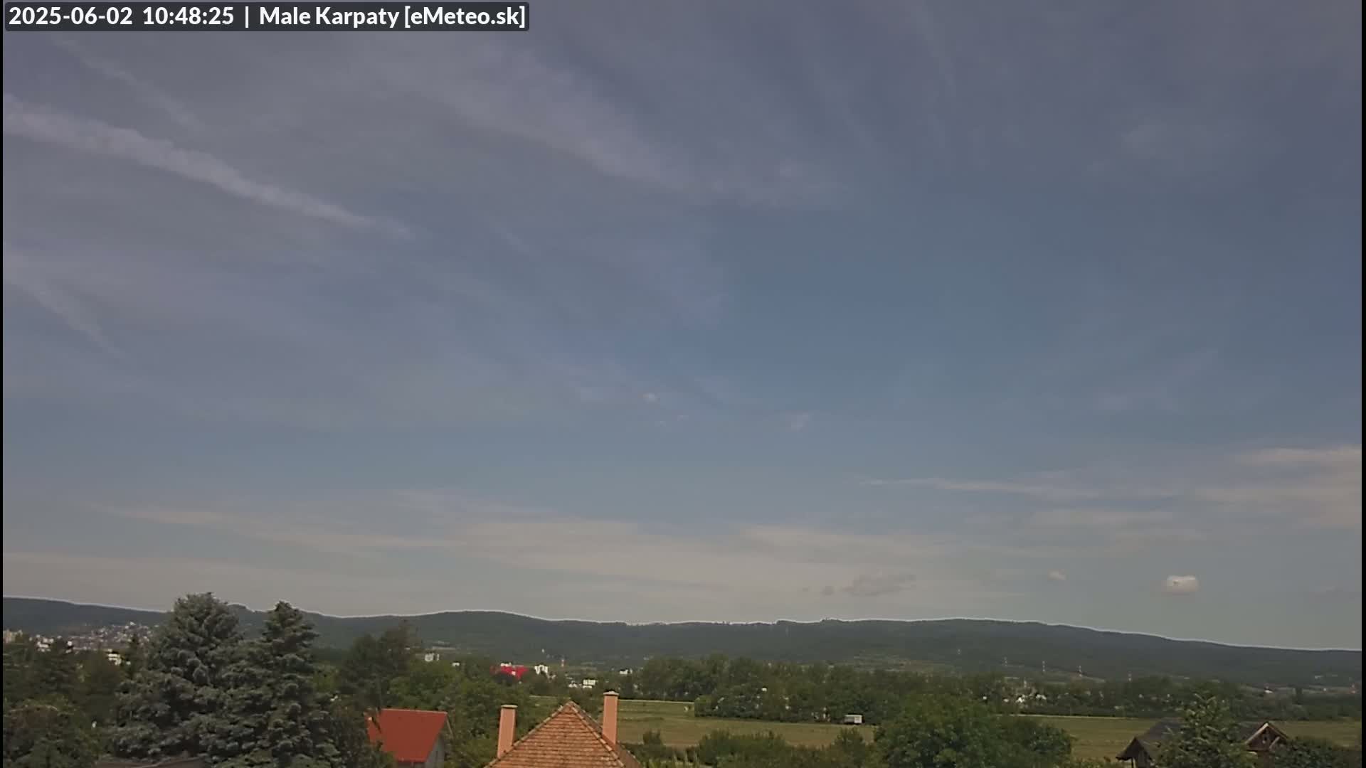 A mostly clear sky with thin, wispy clouds over a landscape of green trees, fields, and distant mountains.