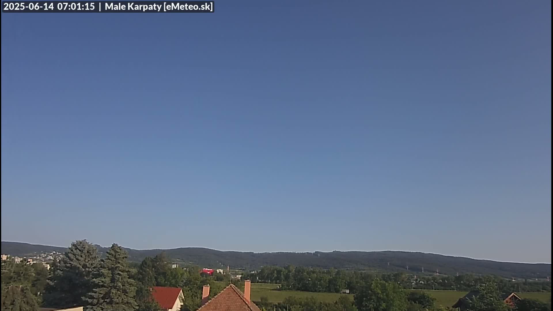 Under a clear blue sky, a range of forested hills stretches across the horizon behind a small town with houses and trees in the foreground.