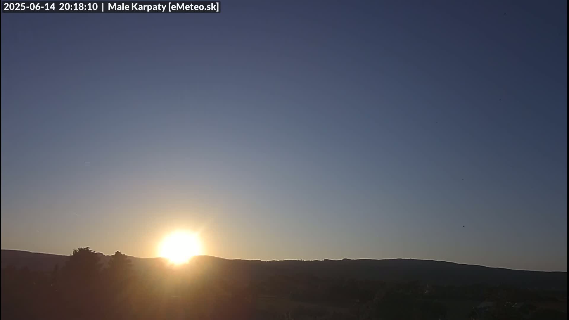 A bright sun sets behind a dark silhouette of low hills and trees under a clear, twilight sky.