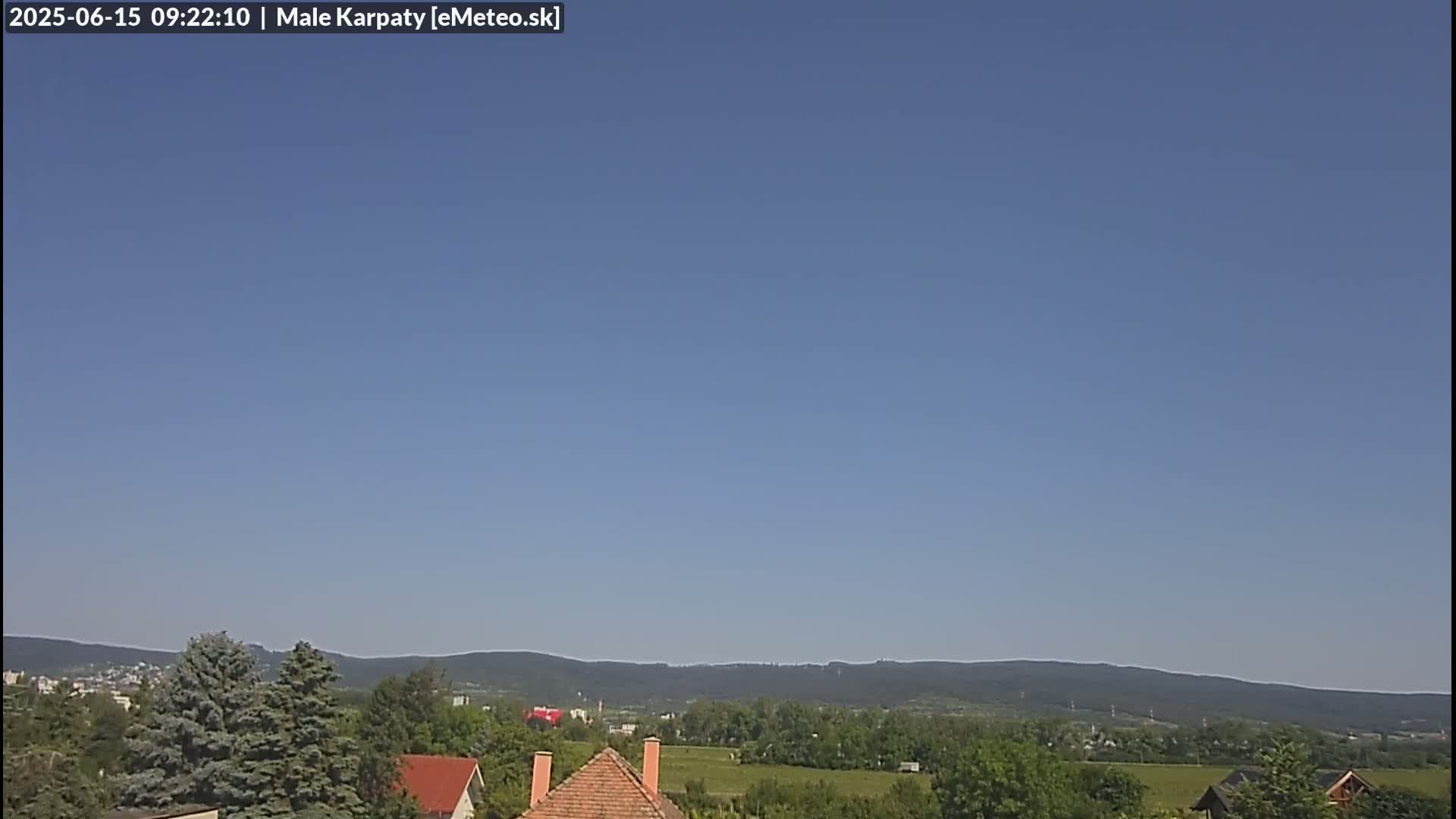 A clear blue sky overlooks a landscape of green trees, fields, and houses with brown roofs, backed by a distant mountain range.