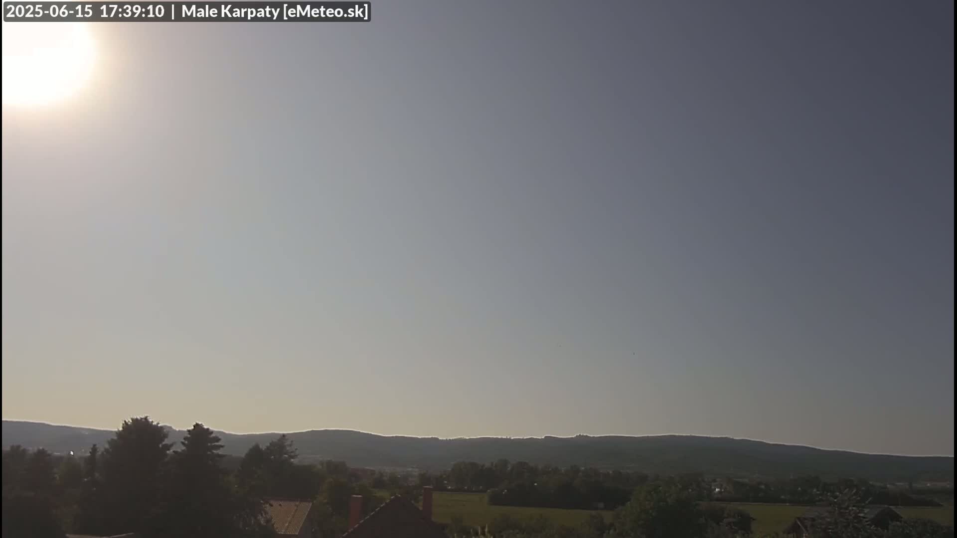 A bright sun shines in a mostly clear sky over a landscape of low-lying hills and green vegetation.