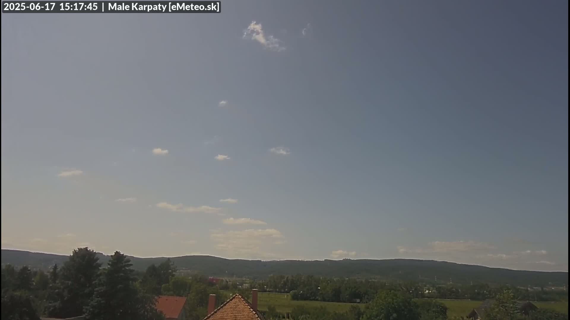 A mostly clear sky with a few scattered clouds overlooks a green landscape of trees, fields, and distant hills.