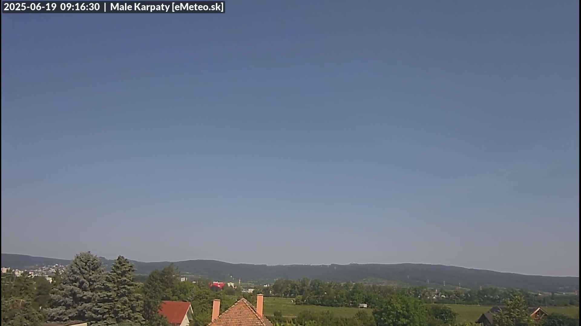A clear, sunny day reveals a distant mountain range behind a line of trees and houses with reddish-brown roofs.