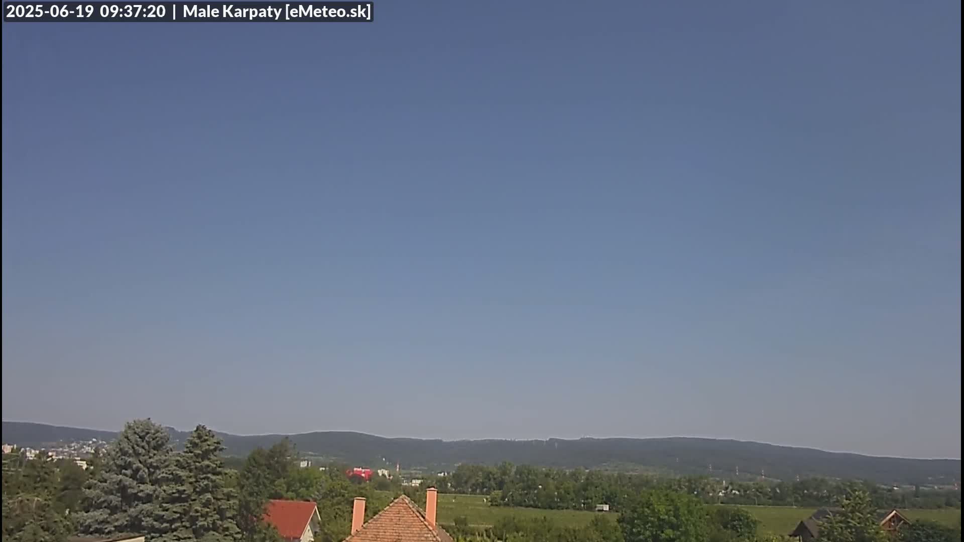 A clear blue sky overlooks a landscape of green trees, distant mountains, and a few houses with red-tiled roofs.