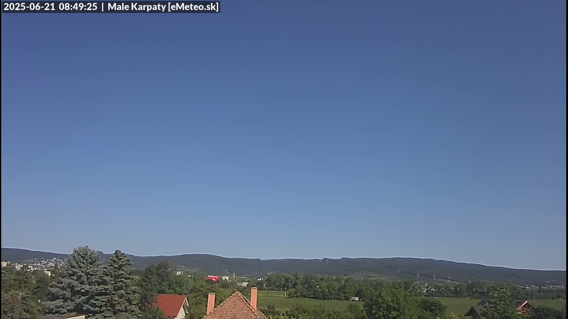 A clear blue sky overlooks a landscape of green trees, fields, and houses with distant mountains under a cloudless sky.