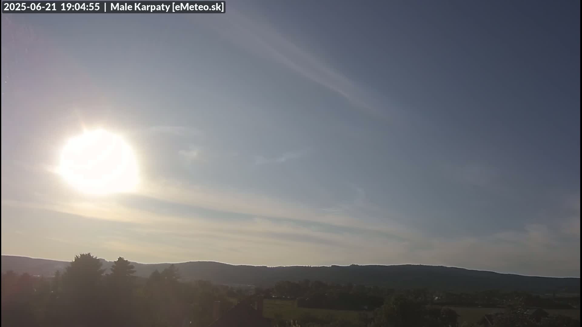 A bright sun shines in a mostly clear sky above a distant line of low hills and a foreground of trees and fields.