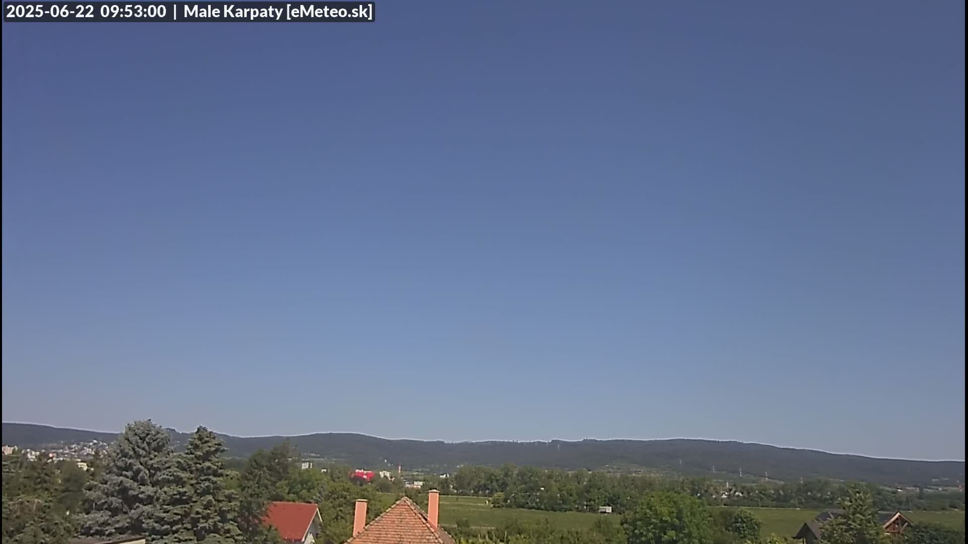 A clear blue sky overlooks a landscape of green trees, fields, and houses with reddish-brown roofs, backed by distant mountains.