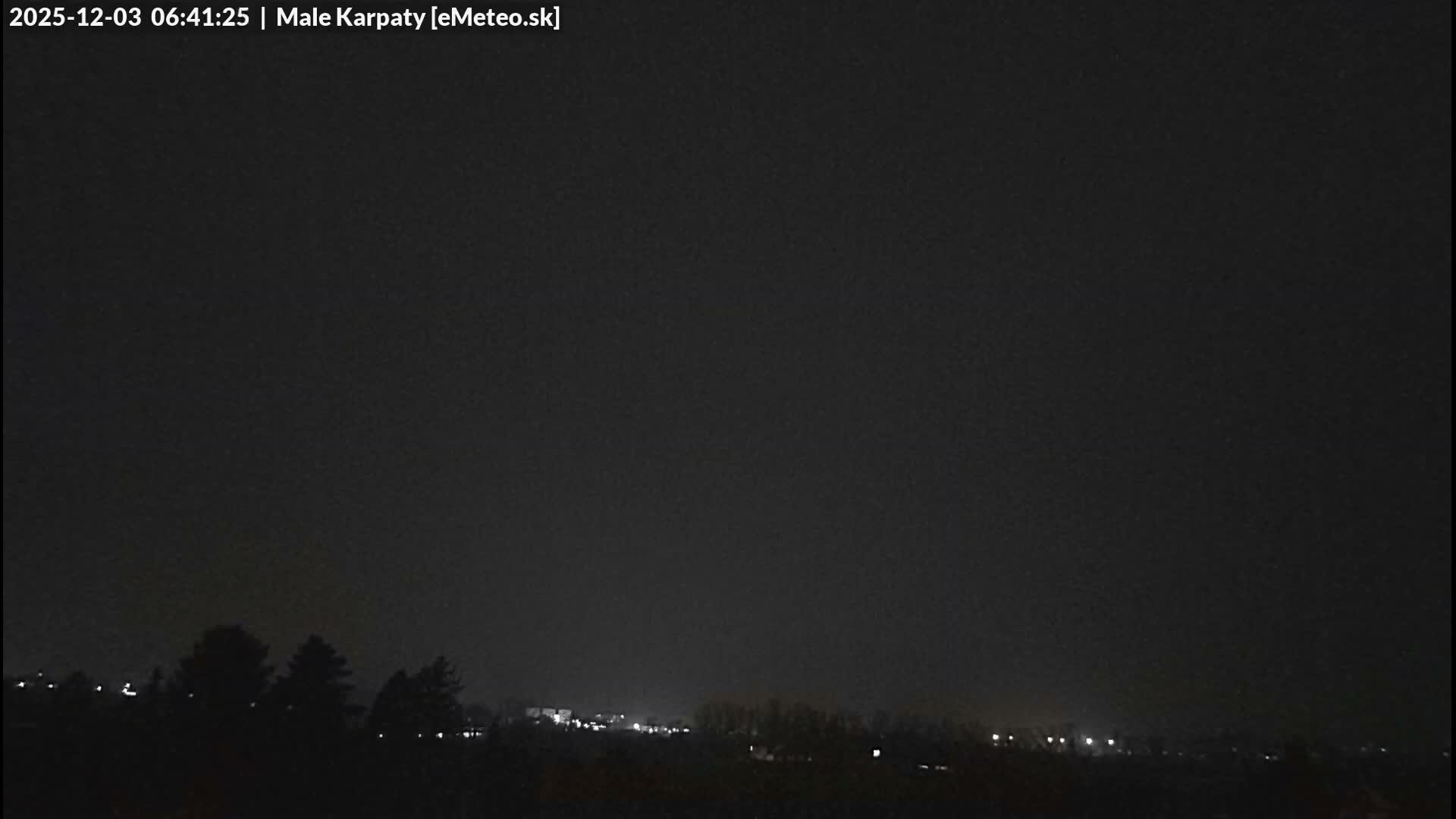 A very dark, early morning outdoor scene reveals the silhouettes of trees on the left and scattered distant lights of a town or village under an overcast sky.