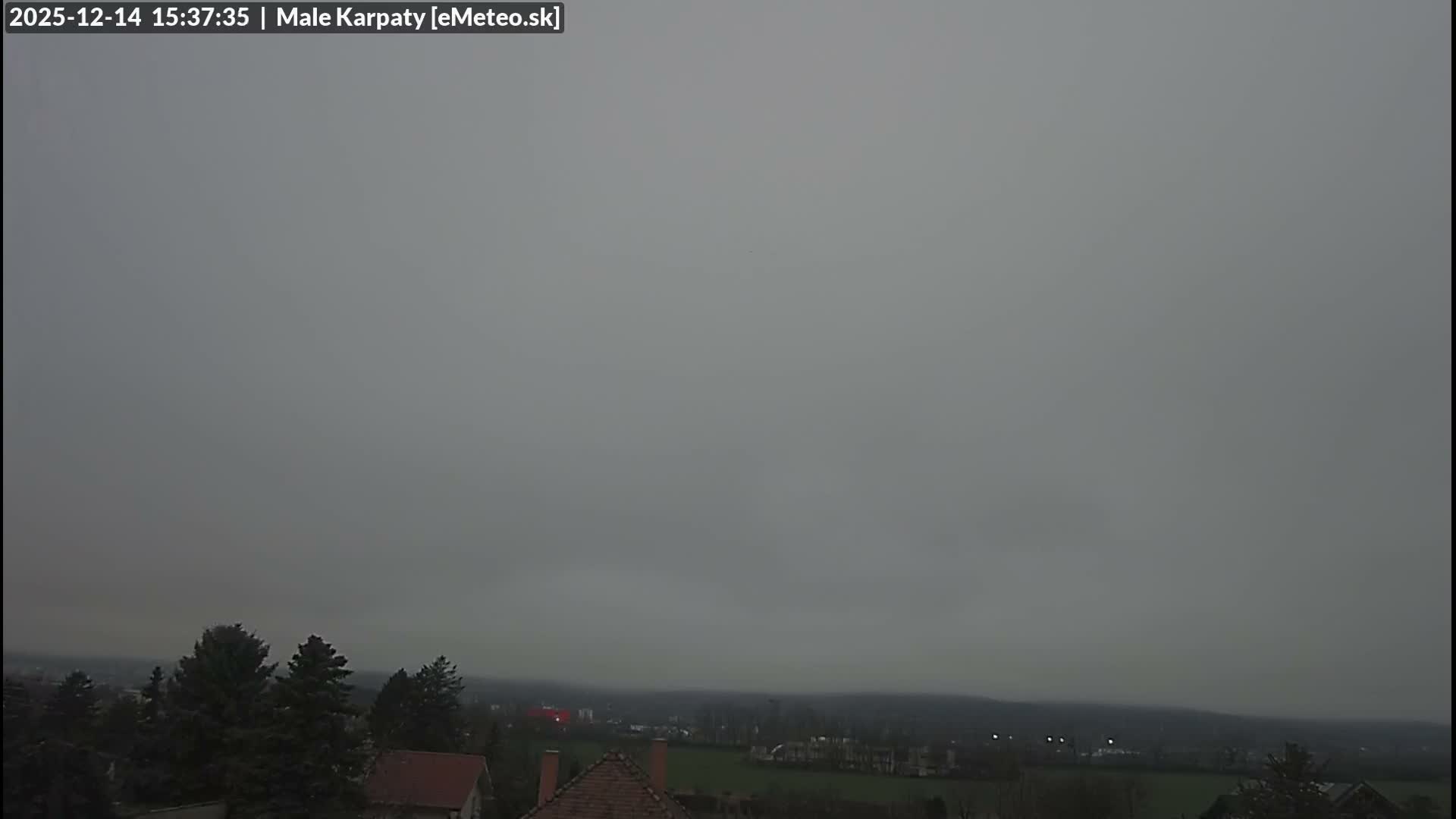 A very dark, early morning outdoor scene reveals the silhouettes of trees on the left and scattered distant lights of a town or village under an overcast sky.