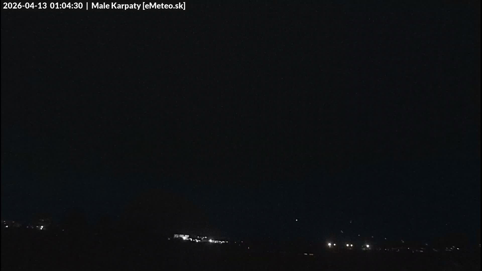 A very dark, early morning outdoor scene reveals the silhouettes of trees on the left and scattered distant lights of a town or village under an overcast sky.