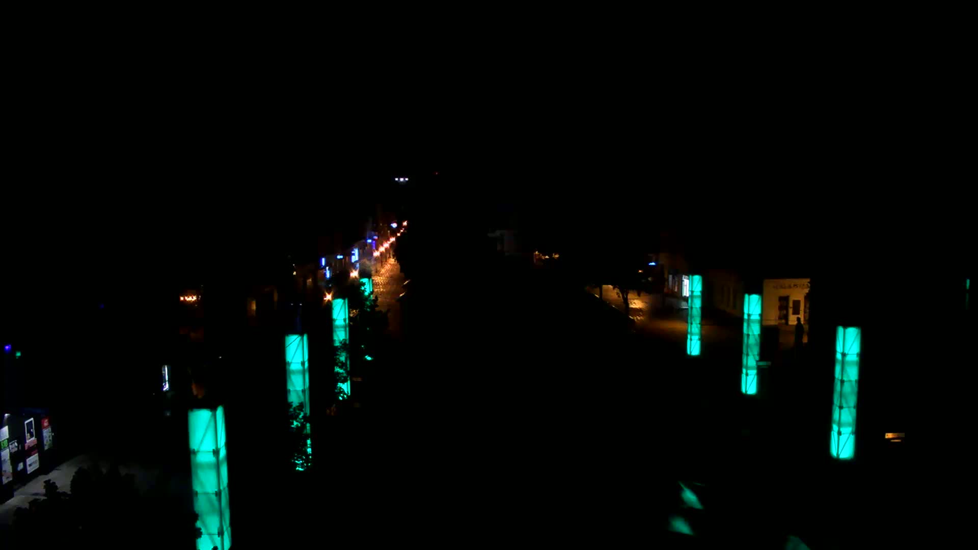 A nighttime view of a road with sparsely spaced vehicles, illuminated by teal-colored pillars lining both sides.