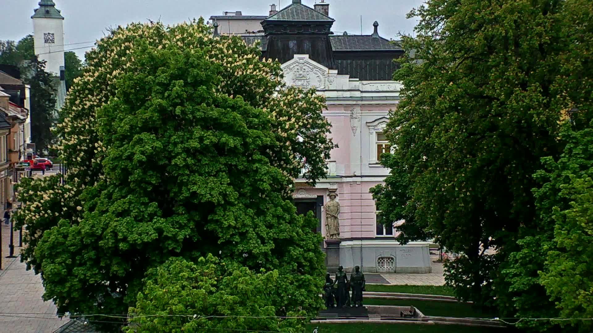 A pink building with ornate details is partially obscured by lush green trees in bloom, with a statue in the foreground on a cloudy day.