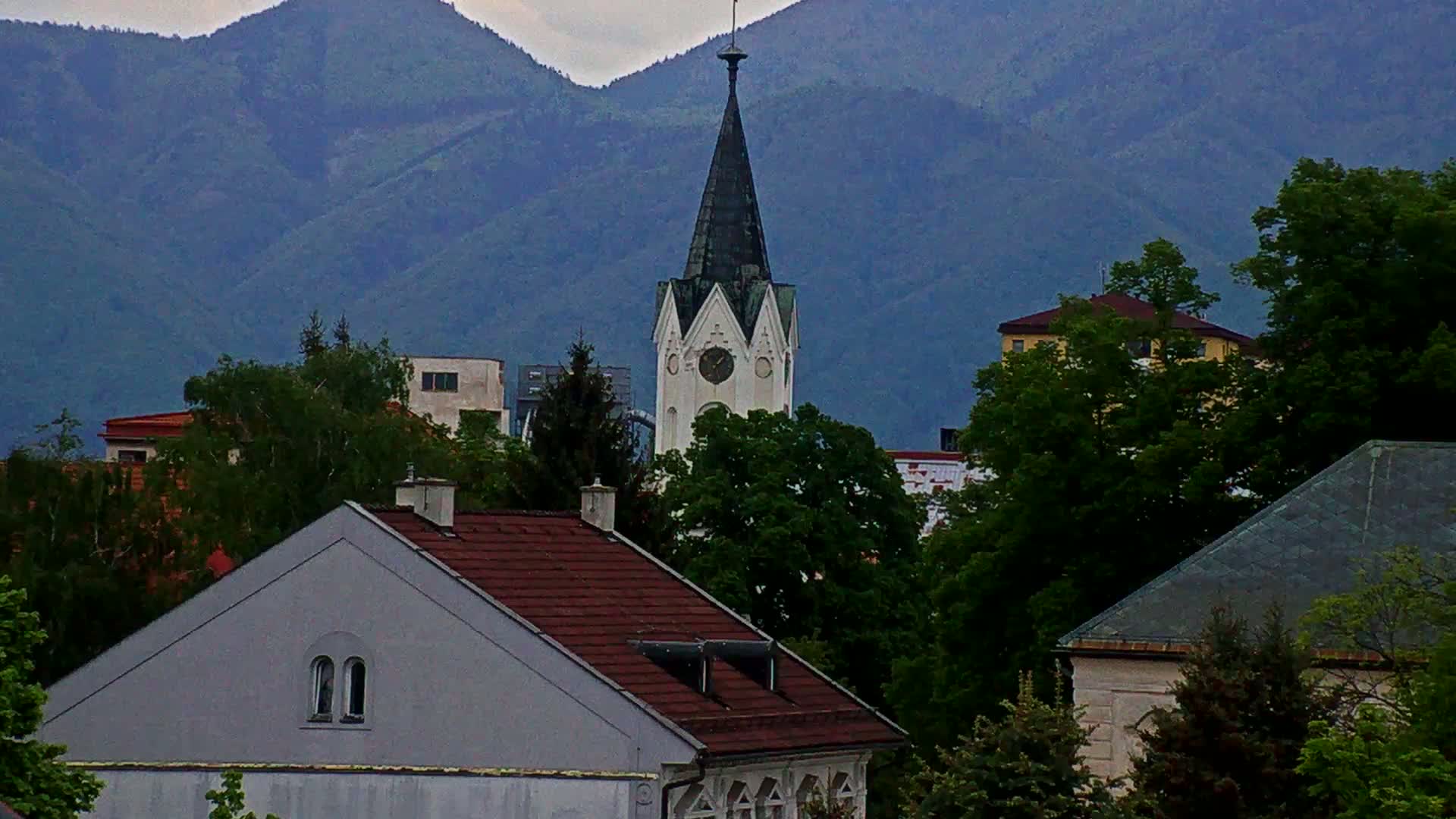 A white church steeple with a clock is visible among a cluster of buildings and lush green trees against a backdrop of overcast mountains.