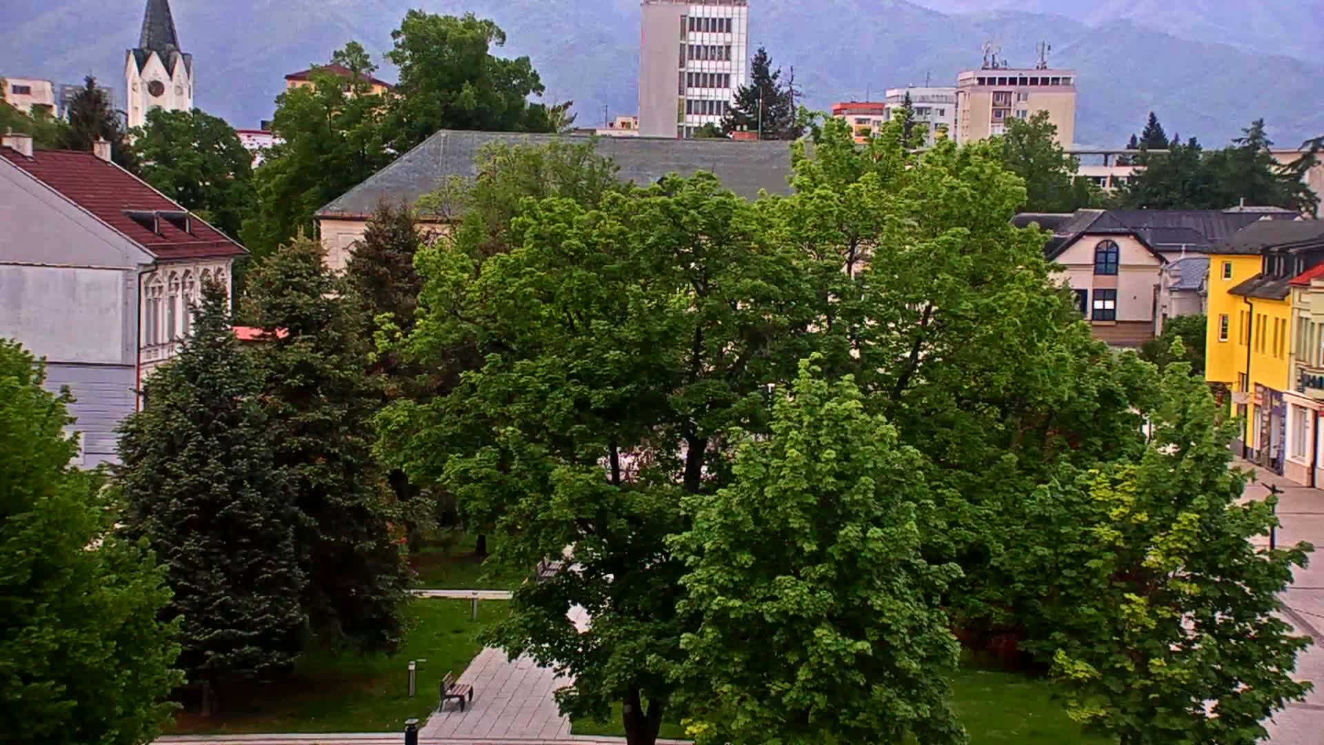 A lush green park sits in the center of a town with various buildings, including a church spire and multi-story buildings, under an overcast sky.