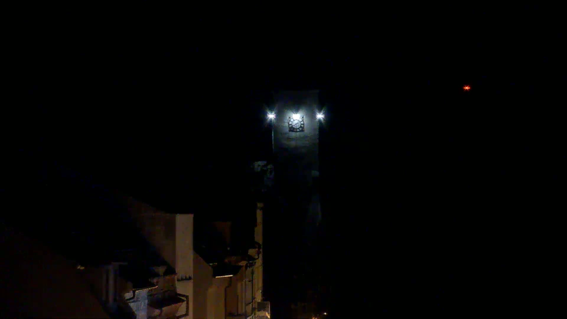 A tall, illuminated clock tower stands against a dark, nighttime sky, with low-lit buildings visible in the foreground.