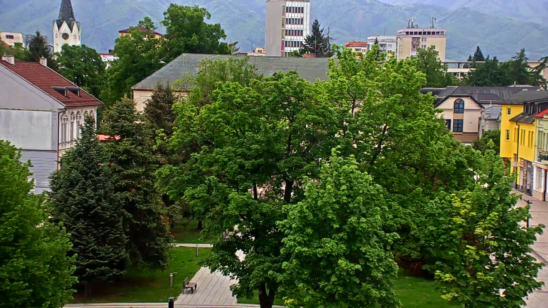 A green park with lush trees sits among buildings in a town, with mountains visible in the background under an overcast sky.
