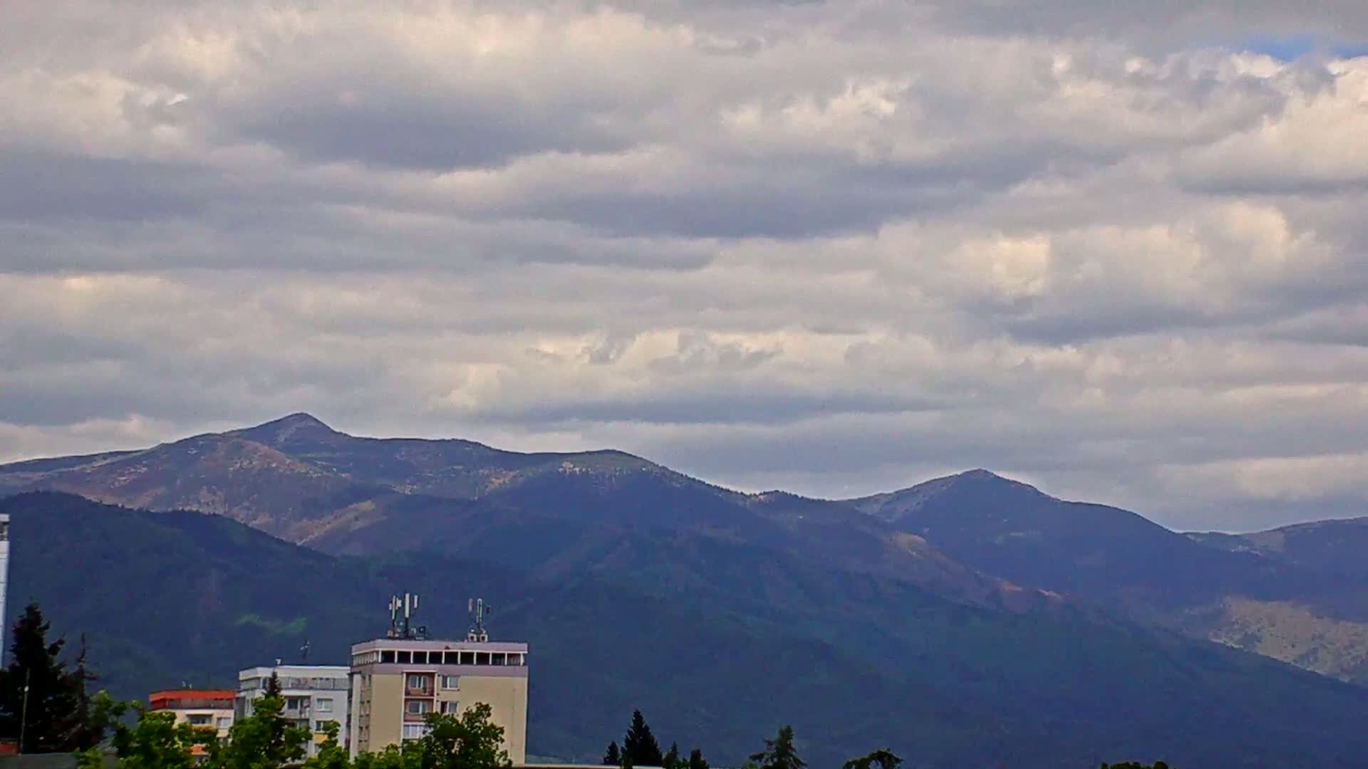 A partly cloudy sky overlooks a range of dark green mountains, behind which sits a multi-story building and some smaller structures.