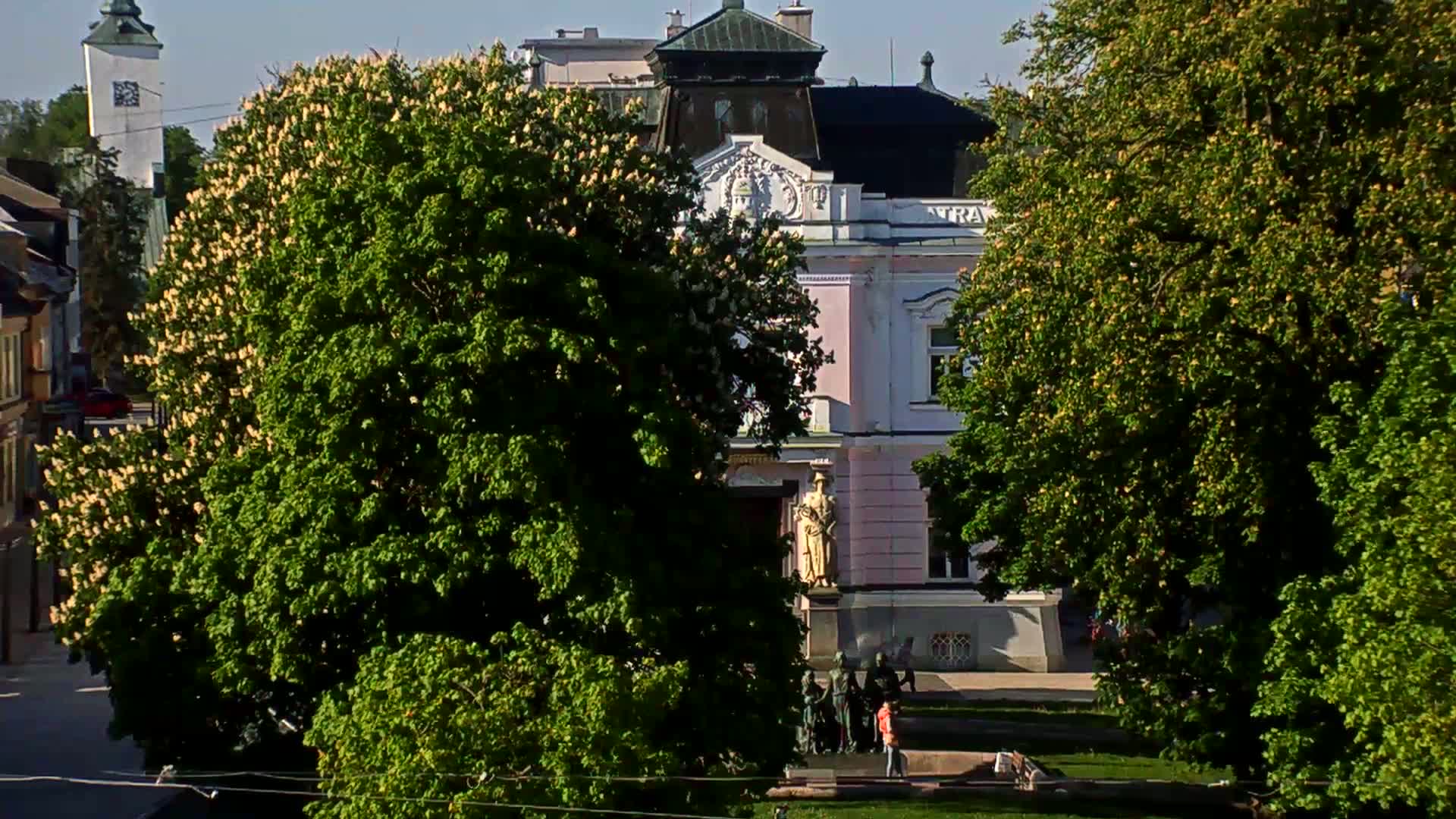 A pink building with a statue in front is partially obscured by lush green trees in bloom on a sunny day.