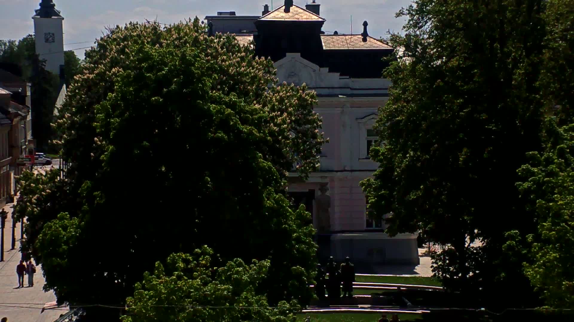 A view of a town square on a sunny day, showing a pink building partially obscured by lush green trees, with a church steeple visible in the distance.