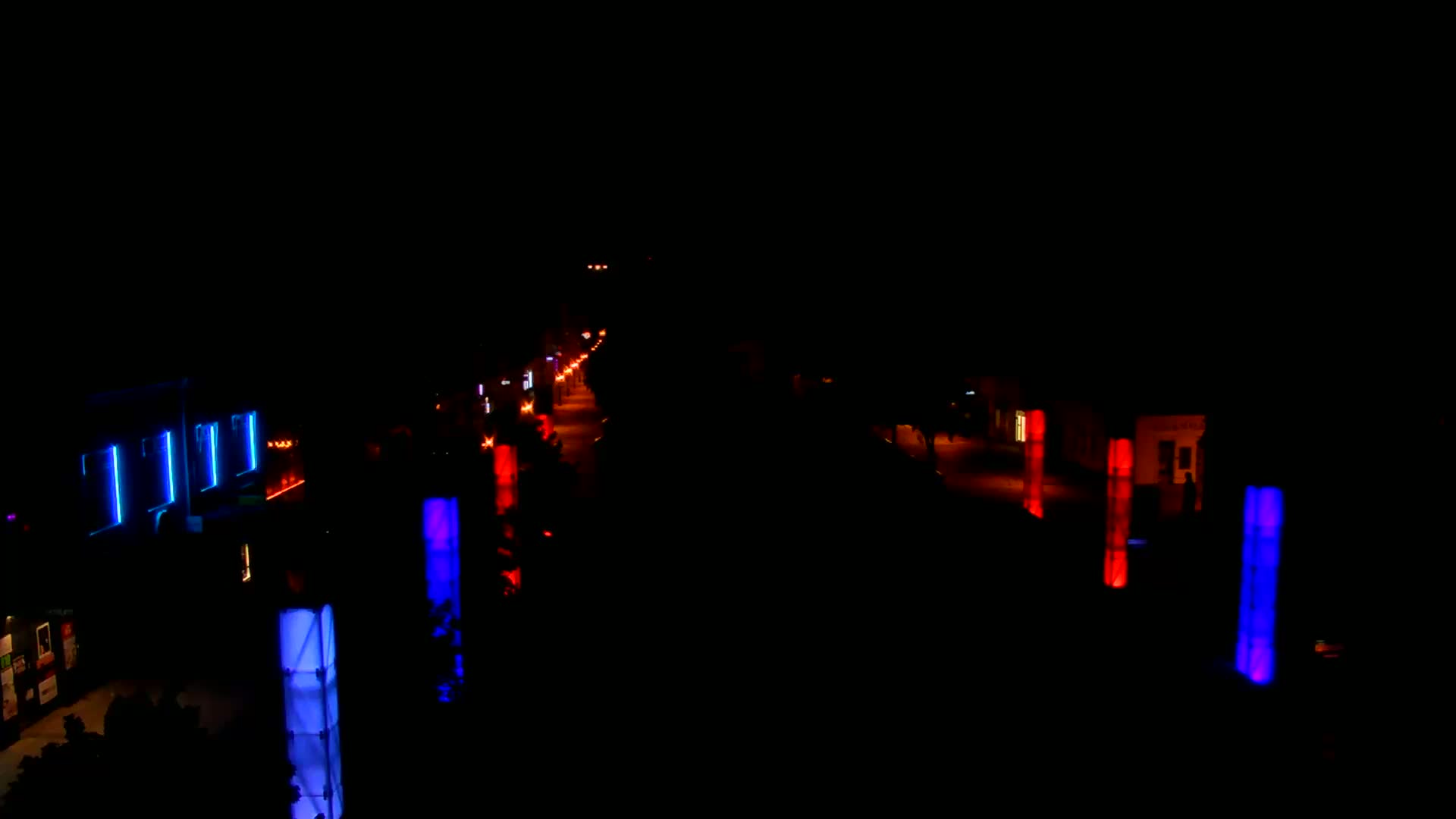 A dark night scene shows a street lit by scattered orange lights and tall, illuminated blue and red pillars along the sides of the road.