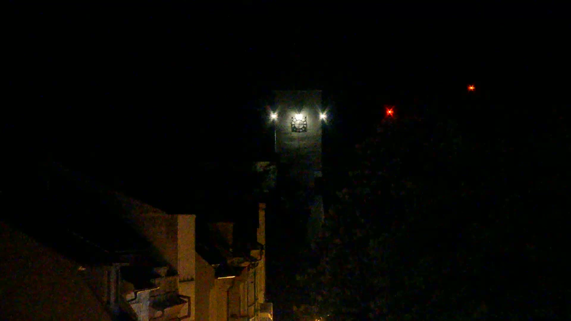 A clock tower is illuminated at night, with two distant red lights visible in the dark sky.