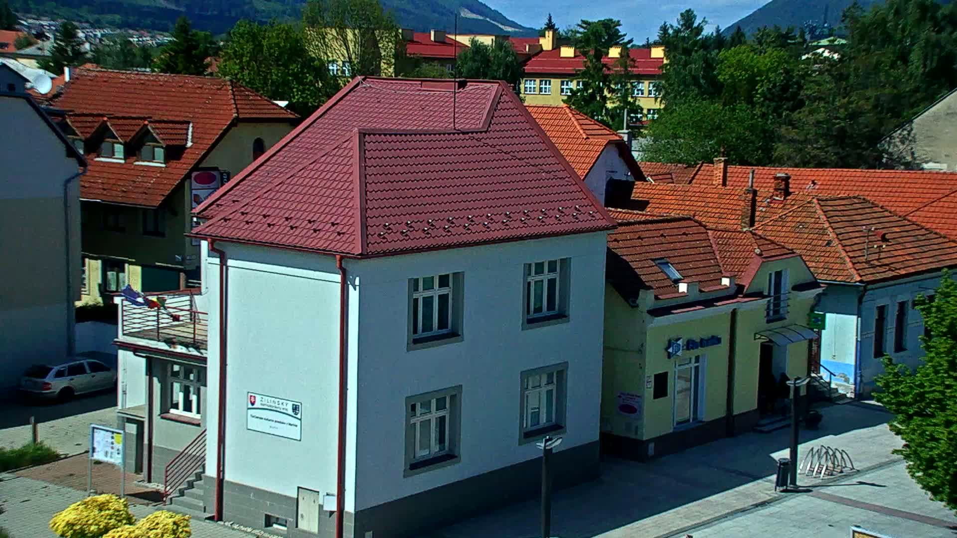 A light-blue two-story building with a reddish-brown roof sits among other buildings with reddish-brown roofs, under a sunny sky, in a town nestled against mountains.
