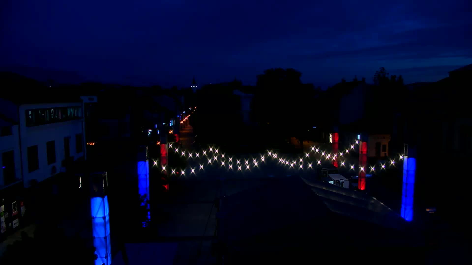 A nighttime street scene shows a string of lights strung across a road between illuminated blue and red pillars, with buildings and trees silhouetted in the background under a dark sky.