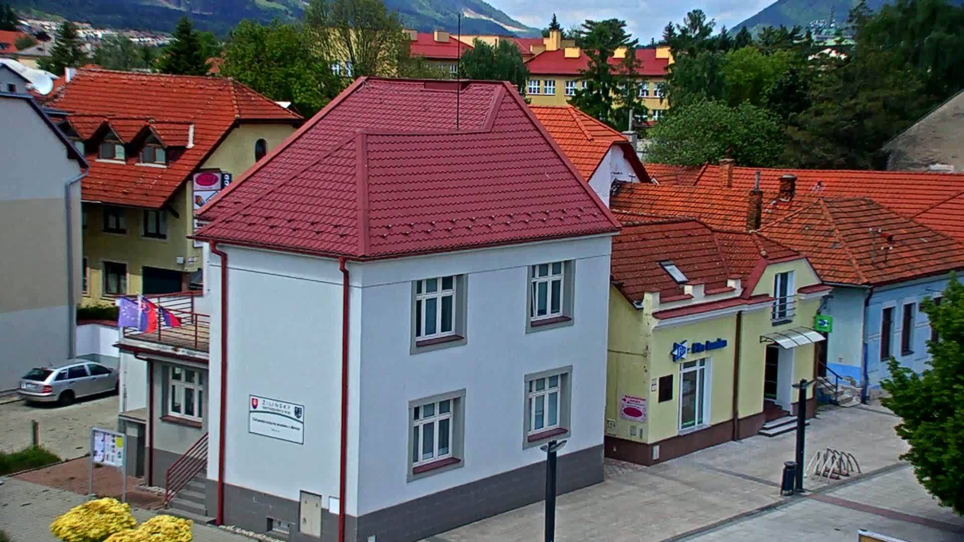 A light-gray building with a red roof sits among other buildings with red tile roofs, all nestled in a town against a backdrop of mountains under a partly cloudy sky.