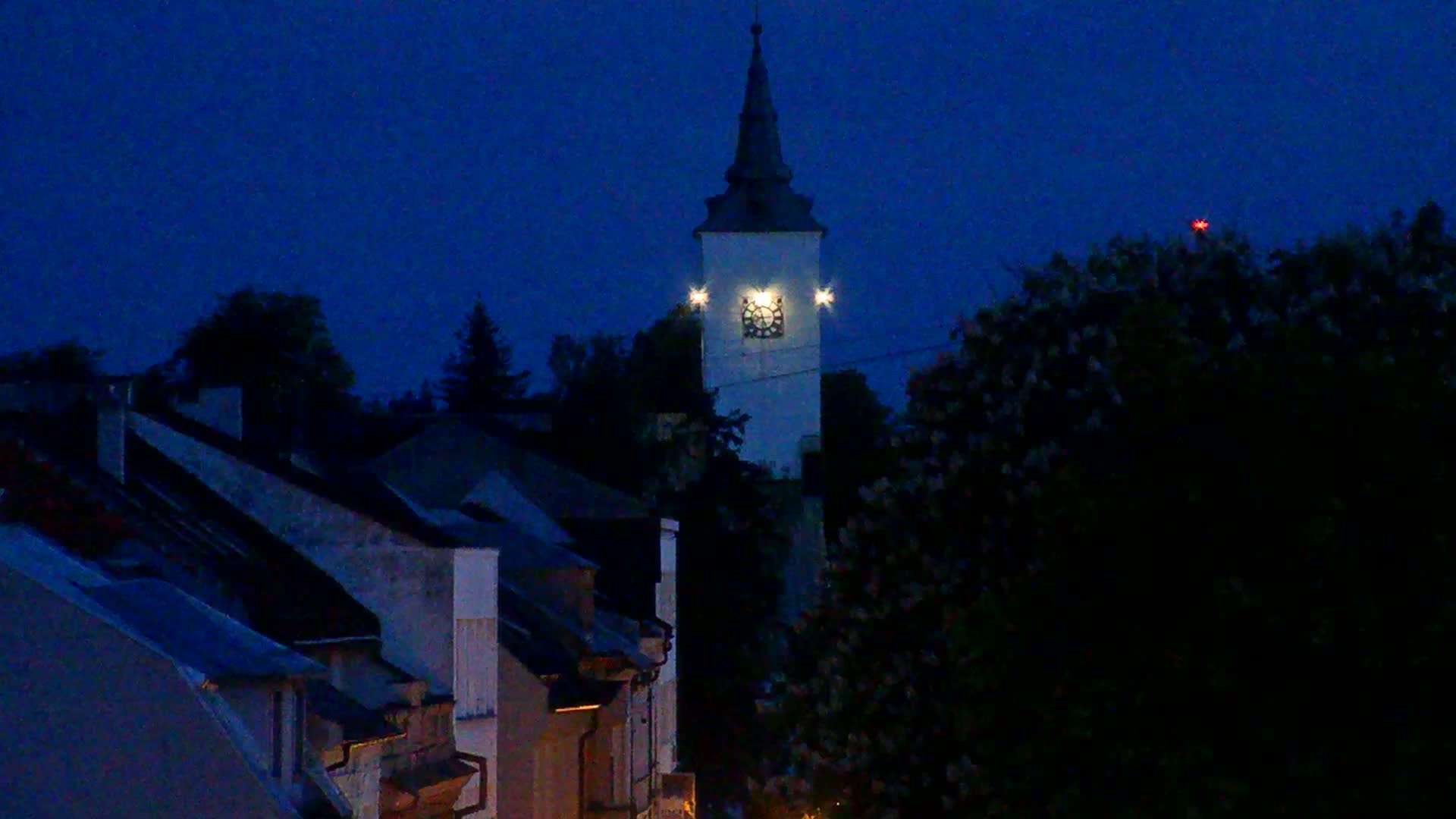 A tall, illuminated clock tower stands above a row of dark houses under a dark blue night sky.