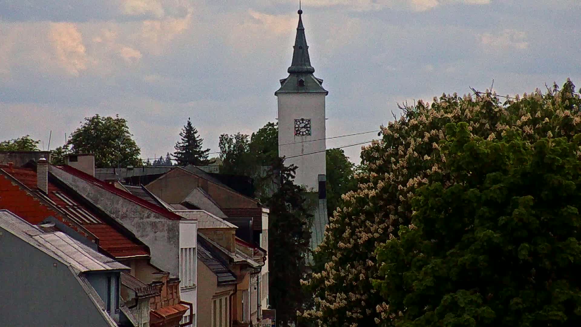 A white tower with a dark spire rises above a cluster of buildings with red-tiled roofs under a cloudy sky.