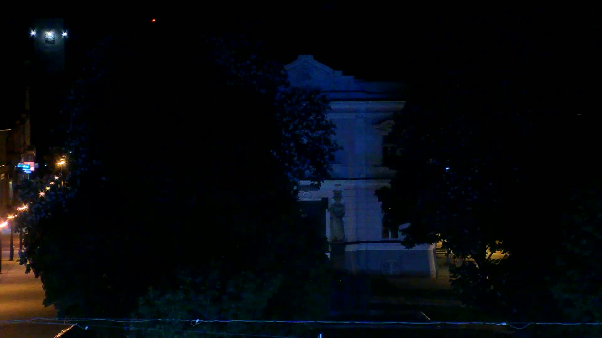 A large building with a statue in front is partially obscured by trees at night under dark conditions.