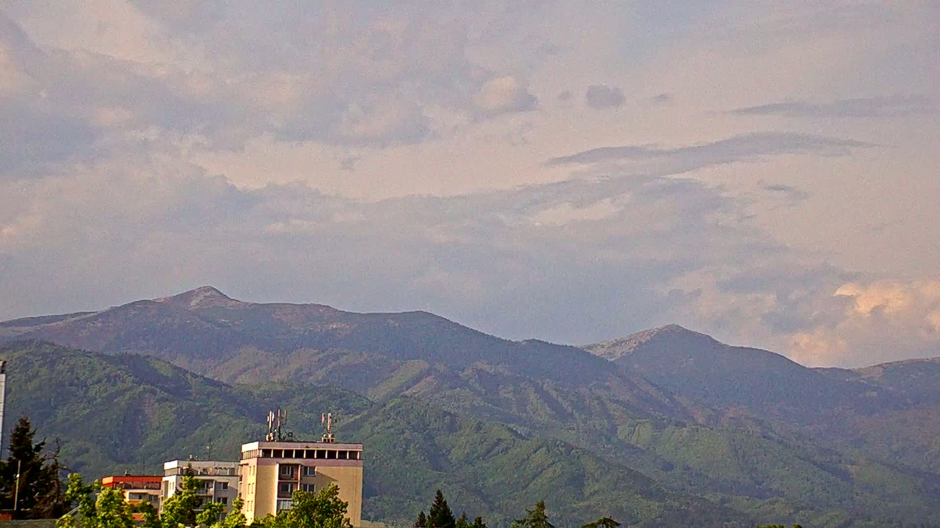 A partly cloudy sky overlooks a range of green-covered mountains, behind which sit several residential buildings.