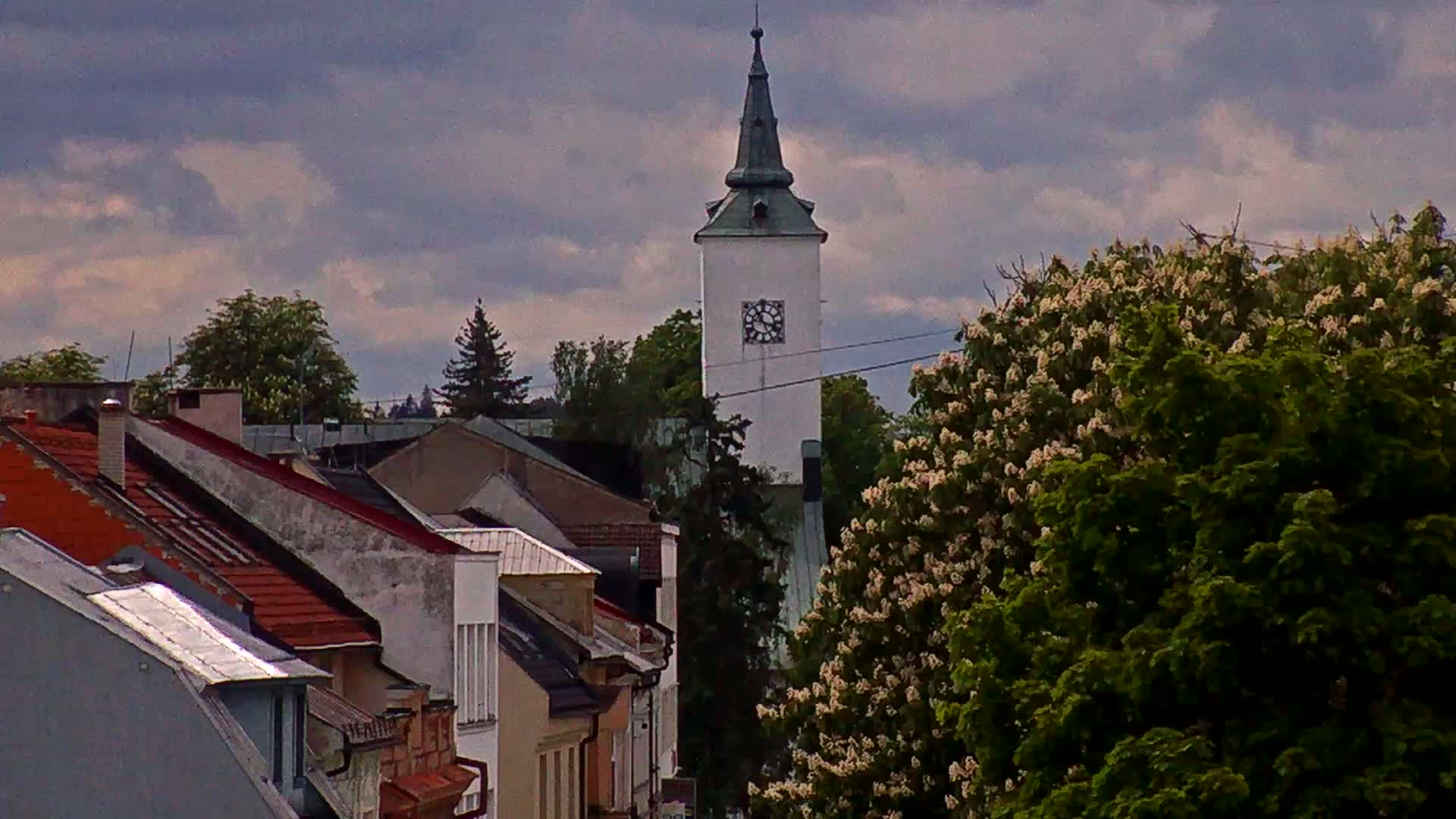 A white clock tower rises above a row of houses with red-tiled roofs under a cloudy sky.