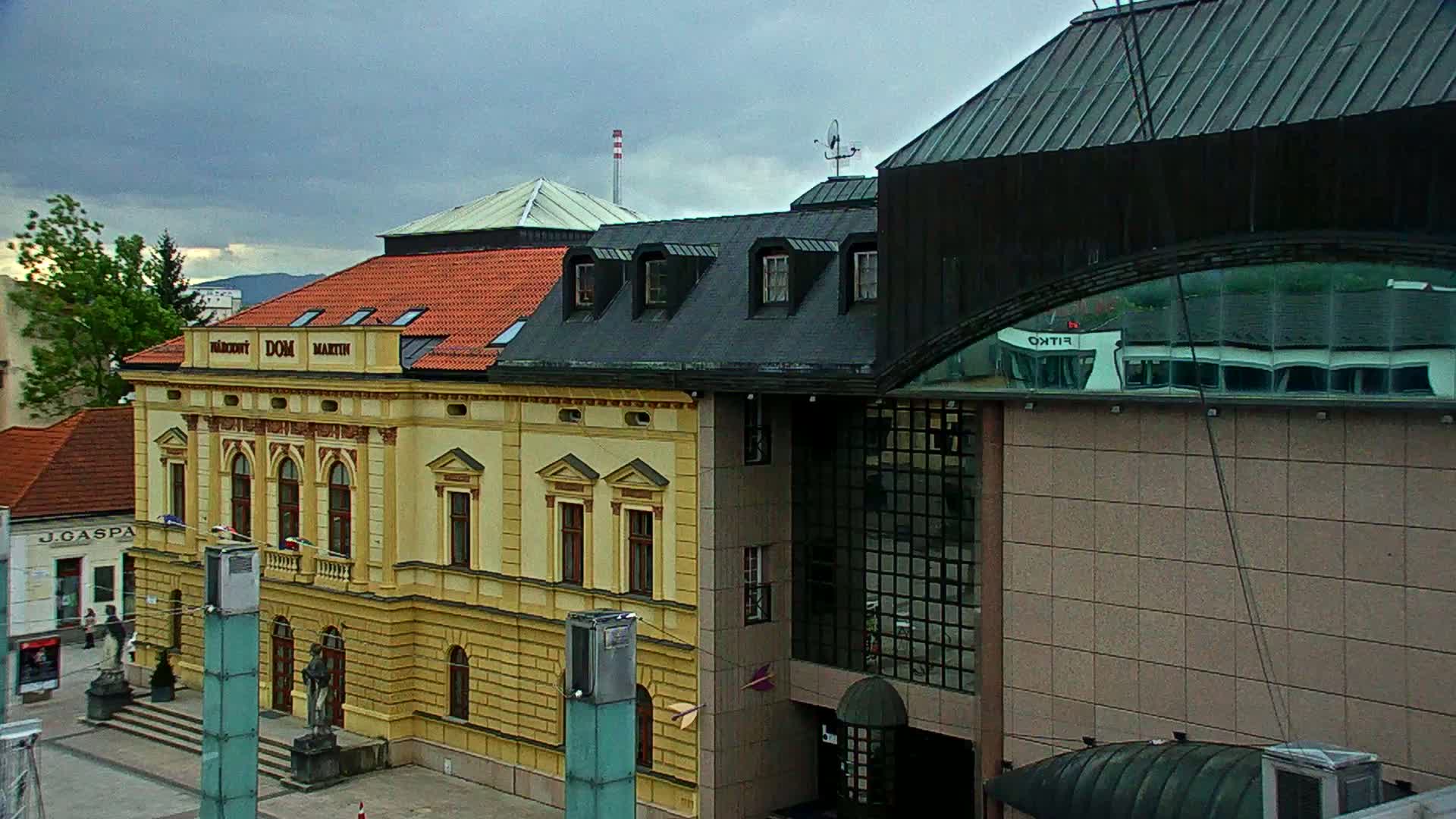 A yellow building with a red roof stands beside a modern, tan building with large windows under an overcast sky.